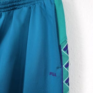 Puede incluir: Pantalones deportivos de color turquesa con una franja lateral turquesa y verde con un patr&oacute;n geom&eacute;trico. Los pantalones tienen una cintura el&aacute;stica y el logotipo "FILA" bordado en morado. Los pantalones est&aacute;n colgados de una percha met&aacute;lica.