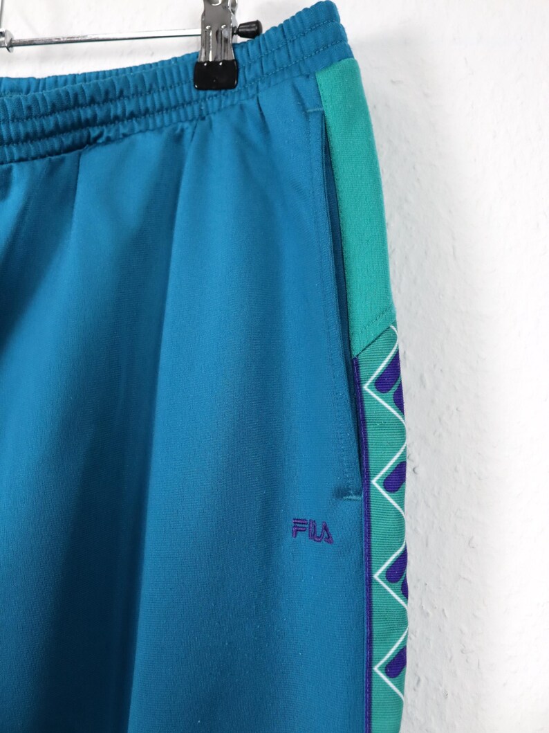 Puede incluir: Pantalones deportivos de color turquesa con una franja lateral turquesa y verde con un patr&oacute;n geom&eacute;trico. Los pantalones tienen una cintura el&aacute;stica y el logotipo "FILA" bordado en morado. Los pantalones est&aacute;n colgados de una percha met&aacute;lica.