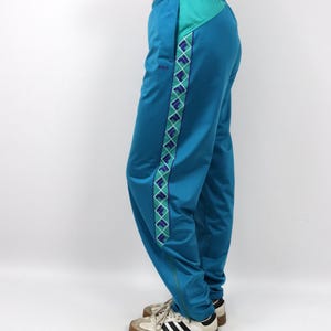 Puede incluir: Pantalones deportivos de color turquesa con un patr&oacute;n de diamantes turquesa y morados en el lateral. Los pantalones tienen un panel turquesa en la parte superior y un peque&ntilde;o logotipo. Se combinan con zapatillas blancas y negras.