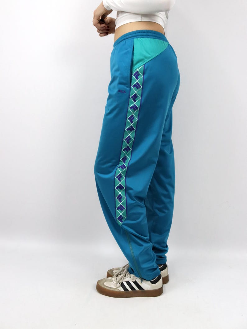 Puede incluir: Pantalones deportivos de color turquesa con un patr&oacute;n de diamantes turquesa y morados en el lateral. Los pantalones tienen un panel turquesa en la parte superior y un peque&ntilde;o logotipo. Se combinan con zapatillas blancas y negras.