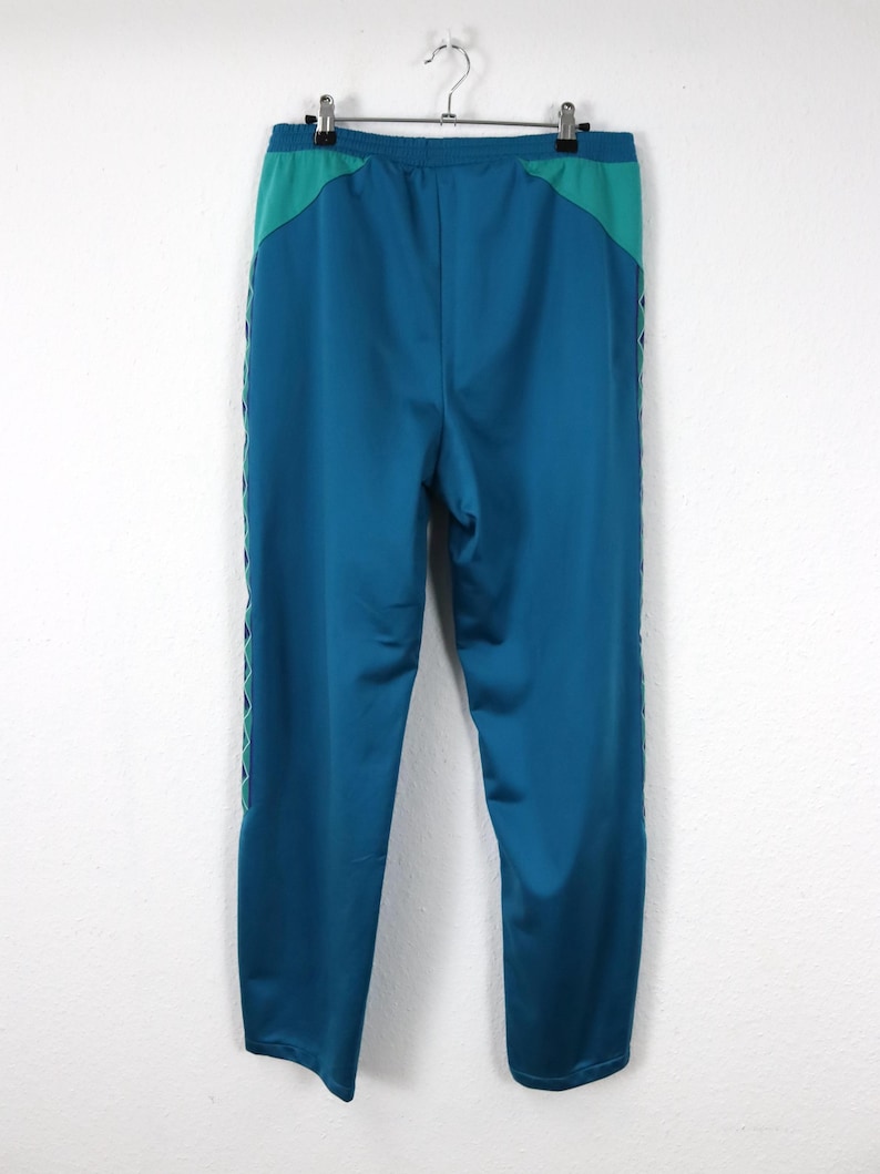 Puede incluir: Pantalones deportivos de color turquesa y turquesa con un panel turquesa en contraste en la cintura y una raya decorativa blanca y turquesa en el lateral. Los pantalones est&aacute;n colgados de una percha met&aacute;lica sobre un fondo blanco.