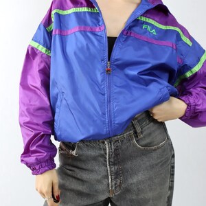 May include: A vintage Fila track jacket in blue, purple, and green. The jacket has a full zipper, a high collar, and the Fila logo on the chest. The sleeves and shoulders are purple, with green and purple stripes across the chest and arms.