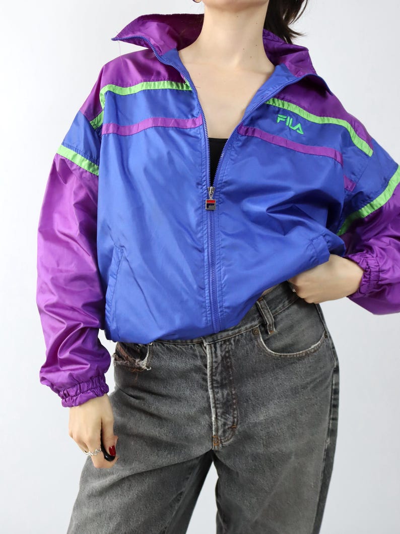 May include: A vintage Fila track jacket in blue, purple, and green. The jacket has a full zipper, a high collar, and the Fila logo on the chest. The sleeves and shoulders are purple, with green and purple stripes across the chest and arms.