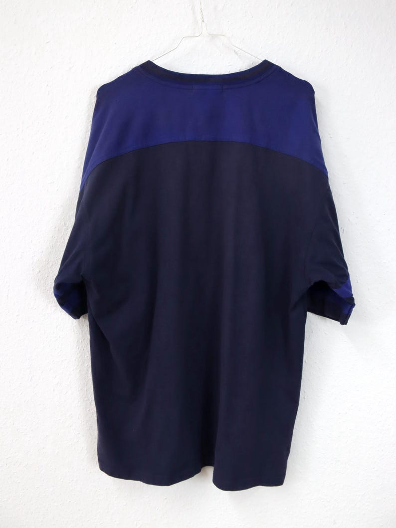 May include: A navy blue and dark blue color-blocked t-shirt hanging on a white hanger. The shirt has a crew neck and short sleeves. The top portion of the shirt is dark blue, and the bottom portion is navy blue.