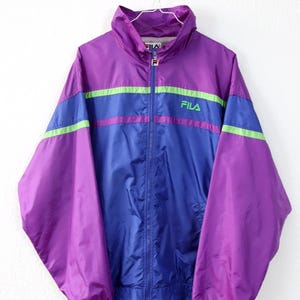May include: A vintage Fila track jacket in purple, blue, and green. The jacket has a high collar, a full zipper, and the Fila logo on the chest. The sleeves and body are a mix of purple and blue with green and purple stripes across the chest.
