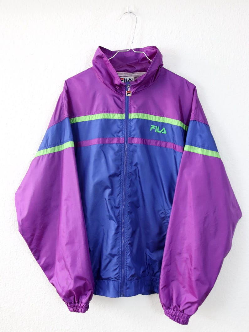 May include: A vintage Fila track jacket in purple, blue, and green. The jacket has a high collar, a full zipper, and the Fila logo on the chest. The sleeves and body are a mix of purple and blue with green and purple stripes across the chest.