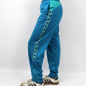 Puede incluir: Pantalones deportivos de color turquesa con un dise&ntilde;o geom&eacute;trico turquesa y blanco en el lateral. Los pantalones tienen un bolsillo turquesa y una cremallera en el tobillo. Los pantalones se combinan con zapatillas blancas y negras.