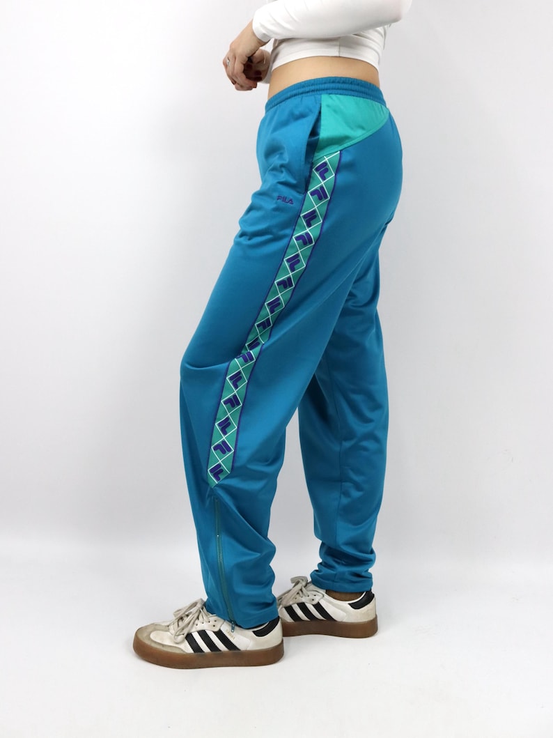 Puede incluir: Pantalones deportivos de color turquesa con un dise&ntilde;o geom&eacute;trico turquesa y blanco en el lateral. Los pantalones tienen un bolsillo turquesa y una cremallera en el tobillo. Los pantalones se combinan con zapatillas blancas y negras.