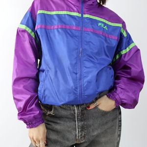 May include: A vintage FILA track jacket in purple, blue, and green. The jacket has a high collar, a full zipper, and the FILA logo on the chest. The sleeves and lower body are purple, with blue and green stripes across the chest.