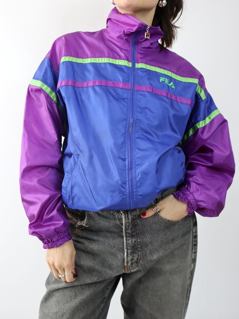 May include: A vintage FILA track jacket in purple, blue, and green. The jacket has a high collar, a full zipper, and the FILA logo on the chest. The sleeves and lower body are purple, with blue and green stripes across the chest.