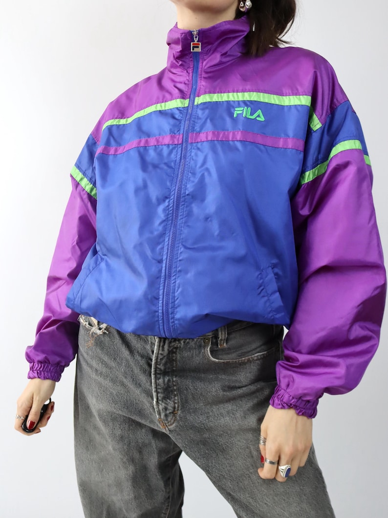 May include: A vintage Fila track jacket in purple, blue, and green. The jacket has a high collar, a full zipper, and the Fila logo on the chest. The sleeves and waist are elasticized.