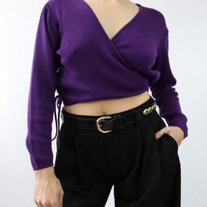 Könnte beinhalten: Ein lila Wickelpullover mit langen Ärmeln und V-Ausschnitt. Der Pullover wird mit einer schwarzen Hose und einem schwarzen Gürtel mit goldener Schnalle kombiniert. Der Gürtel hat goldene Kettendetails. Das Outfit ist schlicht und elegant.