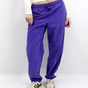 Può includere: Una persona indossa una maglia bianca a maniche lunghe e pantaloni della tuta viola con coulisse. La persona indossa anche scarpe da ginnastica bianche e beige con strisce nere.