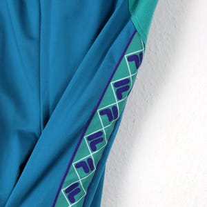 Puede incluir: Pantalones deportivos de color turquesa con una franja verde y morada. La franja presenta un patr&oacute;n de logotipo repetido. Los pantalones tienen una cintura con cord&oacute;n y una combinaci&oacute;n de colores turquesa. Los pantalones est&aacute;n hechos de un material liso, posiblemente sint&eacute;tico.