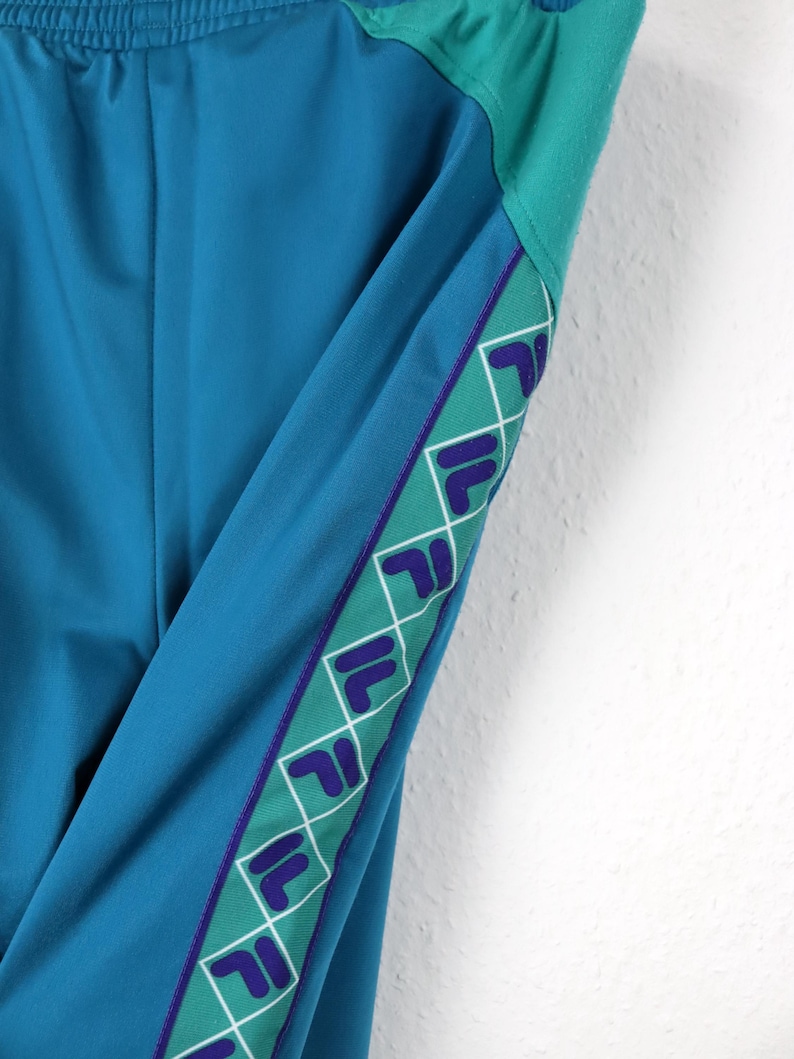 Puede incluir: Pantalones deportivos de color turquesa con una franja verde y morada. La franja presenta un patr&oacute;n de logotipo repetido. Los pantalones tienen una cintura con cord&oacute;n y una combinaci&oacute;n de colores turquesa. Los pantalones est&aacute;n hechos de un material liso, posiblemente sint&eacute;tico.