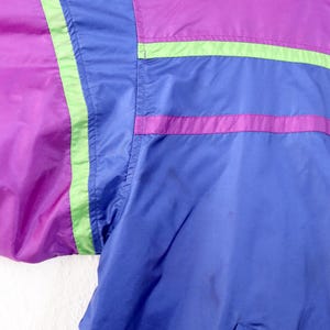May include: Close-up of a vintage jacket in shades of purple, blue, and green. The jacket features a color-block design with horizontal stripes and a pocket. The material appears to be a lightweight nylon or similar fabric.