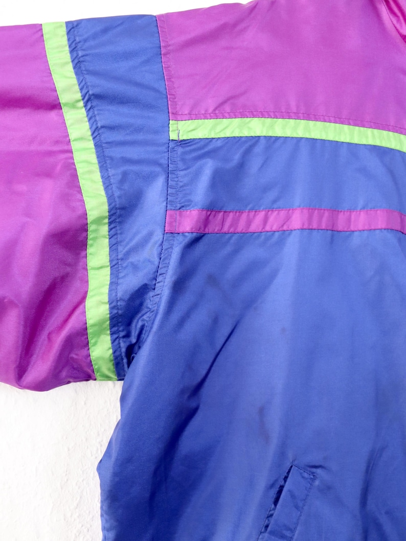 May include: Close-up of a vintage jacket in shades of purple, blue, and green. The jacket features a color-block design with horizontal stripes and a pocket. The material appears to be a lightweight nylon or similar fabric.