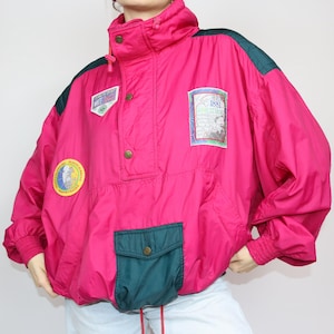 Könnte beinhalten: Eine rosafarbene, grüne und weiße Windbreaker-Jacke mit Reißverschluss und Druckknopfverschluss. Die Jacke hat eine große Tasche auf der Vorderseite und mehrere Patches mit Text und Grafiken. Der Text auf den Patches lautet "White Magic" und "1981 World Championships".