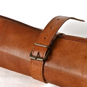May include: A rolled-up brown leather case with a buckle closure. The case is made of a thick, durable leather and has a strap for carrying.