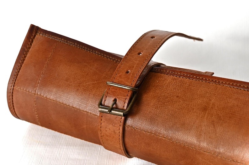 May include: A rolled-up brown leather case with a buckle closure. The case is made of a thick, durable leather and has a strap for carrying.
