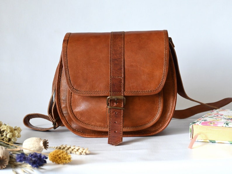 Handgefertigte Umhängetasche aus 100% Naturleder. Halbrunde From mit Druckschnallen-Verschluss in Vintage Optik. Tragriemen in der Länge verstellbar. Frontansicht vor weißem Hintergrund mit Deko