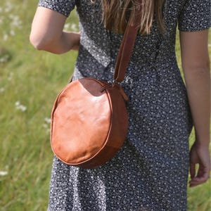 May include: A brown leather round crossbody bag with a long strap. The bag is being worn by a person wearing a floral dress.