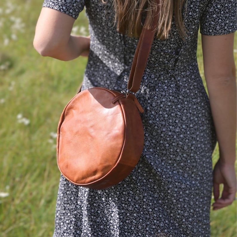 May include: A brown leather round crossbody bag with a long strap. The bag is being worn by a person wearing a floral dress.
