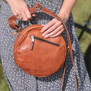 Handgenähte runde Tasche aus natürlich gegerbtem Leder. Trageriemen der Umhängetasche ist der Länge verstellbar.
Nahaufnahme der Tasche in der Hand eines Models.