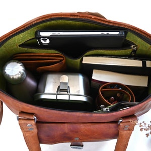 May include: A brown leather tote bag with a green lining. The bag is open and filled with various items, including a tablet, books, a water bottle, a lunchbox, and a pencil case.