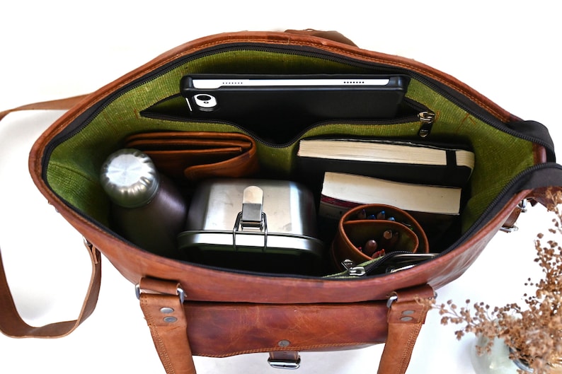 May include: A brown leather tote bag with a green lining. The bag is open and filled with various items, including a tablet, books, a water bottle, a lunchbox, and a pencil case.