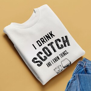 Könnte beinhalten: Weißes Sweatshirt mit dem schwarzen Text "I DRINK SCOTCH AND I KNOW THINGS". Das Design beinhaltet eine Grafik eines Scotch-Glases. Eine Jeans ist in der unteren rechten Ecke zu sehen.