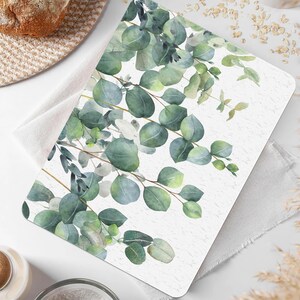 Könnte beinhalten: Ein weißer Platzdeckchen mit einem Aquarell-Design aus grünen Eukalyptusblättern. Das Platzdeckchen hat eine dezente Textur und einen weißen Hintergrund.
