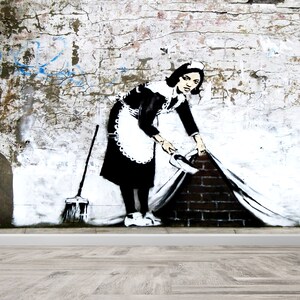 May include: A black and white stencil of a woman in a maid's uniform sweeping a brick wall. The woman is holding a sheet in her right hand and is pulling it away from the wall. The sheet is covering a brick wall. The stencil is on a white brick wall.
