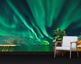 May include: A green and blue aurora borealis, also known as the Northern Lights, shines brightly in the night sky over a snow-covered mountain range and a body of water. A potted plant and a white armchair are in the foreground.