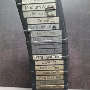 Peut inclure: Un chargeur de fusil noir présentant une série d'échantillons de couleurs horizontaux étiquetés avec du texte incluant White, Medium White, White Gray, Cream, Very Light Gray, Light Gray, Medium Gray, Dark Gray, Very Dark Gray, Very Light Tan, Light Tan, Medium Tan, Dark Tan, Very Dark Tan, Light Brown, Medium Brown et Dark Brown, avec 5.56x45 imprimé en haut.