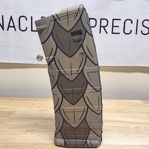 May include: A magazine with a scale pattern in shades of brown and gray. The magazine has the text "5.56 x 45" and "M35" printed on it. The magazine is standing on a wooden surface.