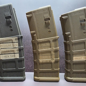 May include: Three polymer rifle magazines in varying shades of tan and dark gray. Each magazine features a textured grid pattern, a rectangular slot, and the text 5.56x45 molded into the top. Labels on the magazines read Light Tan, Medium Tan, Dark Tan, Hex Tan, and Bright Tan.