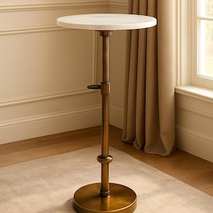 May include: A side table with a round, white marble top and an adjustable brass-coloured metal base. The table has a circular base and a lever for height adjustment. The table is set against a neutral-toned wall and carpet.