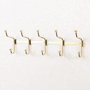 Könnte beinhalten: Ein goldfarbener Wandgarderobenständer mit fünf Doppelhaken. Der Ständer hat eine horizontale Stange mit fünf Hakenpaaren, jeweils mit gebogenem Oberteil und unterem Haken. Das Design ist schlicht und elegant, geeignet für Flure oder Schlafzimmer.