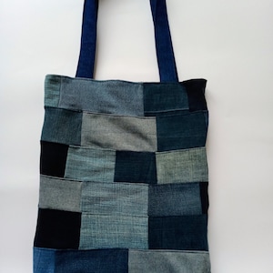 Op de afbeelding: Een blauwe denim tote bag gemaakt van patchwork vierkanten in verschillende tinten blauw denim. De tas heeft twee lange blauwe denim banden.