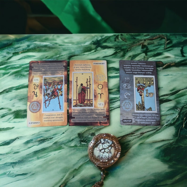 K&ouml;nnte beinhalten: Drei Tarotkarten mit Illustrationen und Text, darunter "CONFLICT" und "FORESIGHT". Ein dekorativer, runder, goldfarbener Anh&auml;nger mit einem baumartigen Design befindet sich im Vordergrund. Der Hintergrund ist eine gr&uuml;ne, marmorierte Oberfl&auml;che.