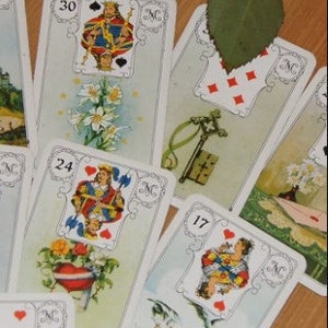 May include: A collection of vintage tarot cards, each featuring detailed illustrations and numbers. The cards are arranged on a wooden surface, with a sprig of greenery in the background. The cards have a white border and are numbered 17, 24, and 30.