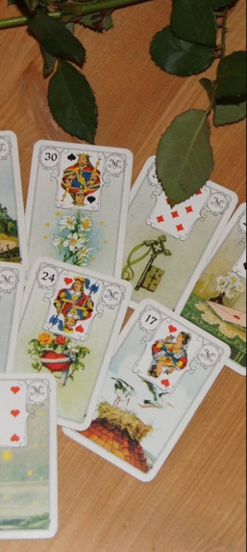 May include: A collection of vintage tarot cards, each featuring detailed illustrations and numbers. The cards are arranged on a wooden surface, with a sprig of greenery in the background. The cards have a white border and are numbered 17, 24, and 30.