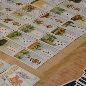 May include: A collection of vintage playing cards, featuring detailed illustrations and traditional suit symbols. The cards are arranged on a wooden surface, with some cards fanned out and others laid out in rows. The image shows a hand pointing at the cards.