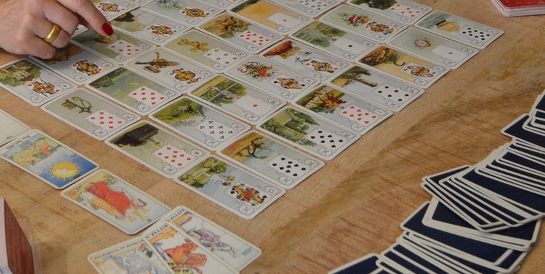 May include: A collection of vintage playing cards, featuring detailed illustrations and traditional suit symbols. The cards are arranged on a wooden surface, with some cards fanned out and others laid out in rows. The image shows a hand pointing at the cards.