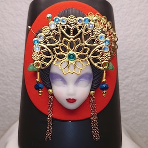 Peut inclure: Mug en céramique noire avec un motif rouge et or représentant un visage de femme avec une coiffe ornée de bijoux.