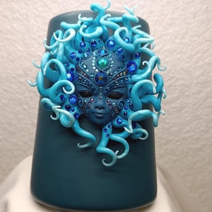 May include: A teal blue ceramic mug with a blue and white sculpted mask with rhinestones. The mask has a third eye and is surrounded by blue and white sculpted tendrils.