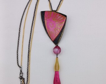Può includere: Una collana con un ciondolo rosa e oro con un fiocco. Il ciondolo è di forma triangolare con un contorno nero e un design rosa e oro. Il fiocco è di colore rosa scuro.