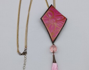 Può includere: Una collana con pendente rosa e oro con un pendente a forma di diamante. Il pendente ha un motivo floreale ed è impreziosito da un nappa rosa e una catena dorata. La collana è lunga circa 50 cm.