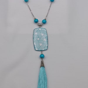 May include: A silver-toned necklace with turquoise beads and a rectangular pendant. The pendant has a white background with a turquoise geometric pattern. A turquoise tassel hangs from the bottom of the pendant. The necklace is a fashion accessory.